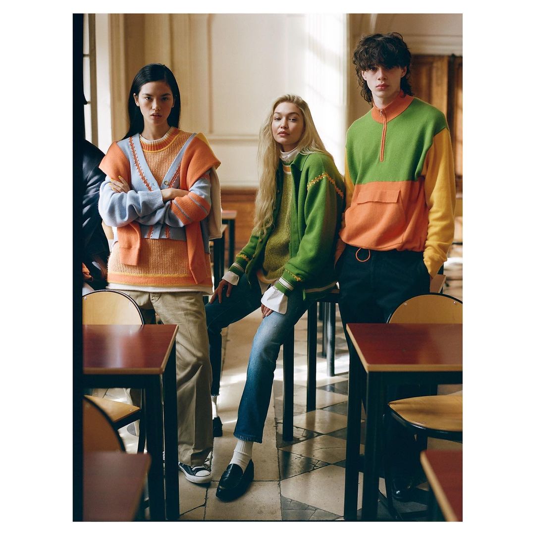 Three fashion models wearing colorful knitwear pose inside a building with tables; a fashion editorial photo.