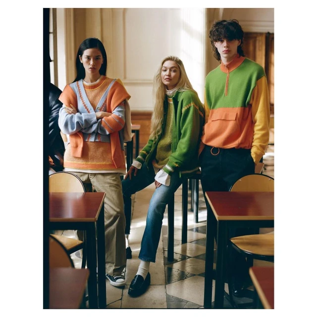 Three fashion models wearing colorful knitwear pose inside a building with tables; a fashion editorial photo.