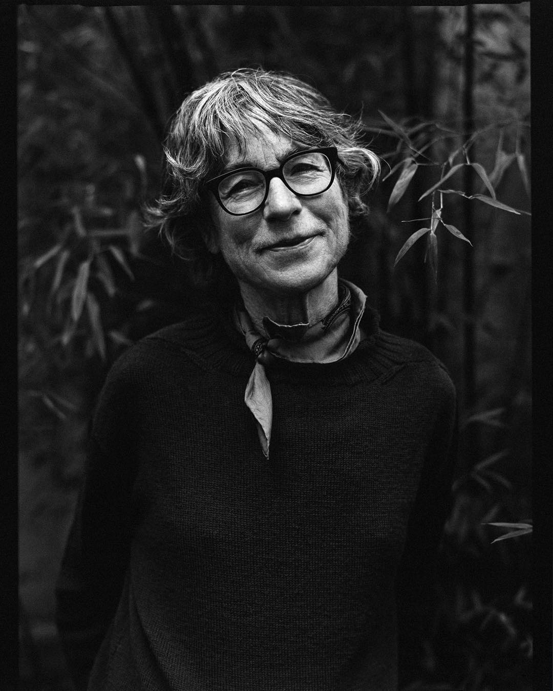 Monochrome portrait of an adult woman wearing glasses and a sweater, set against a lush green backdrop of bamboo and leaves.