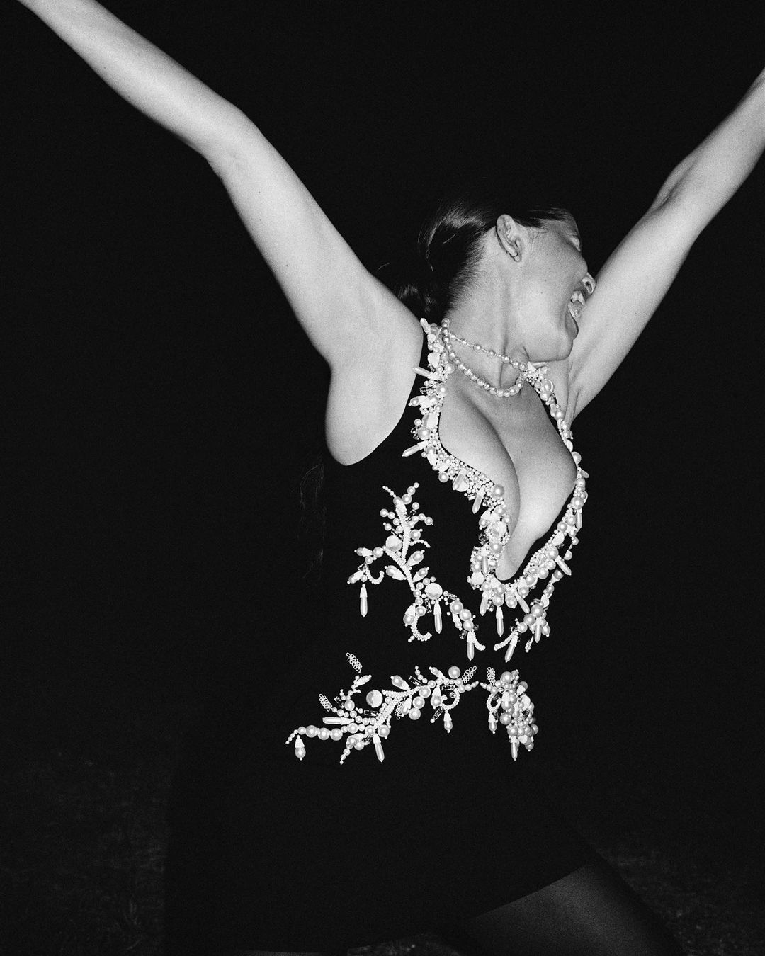 Black and white portrait of a woman with arms raised wearing a black dress adorned with jewels, pearls, and beads, against a dark backdrop.