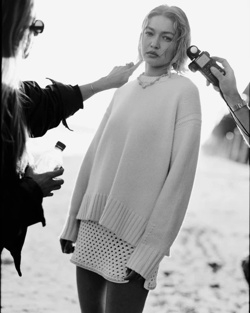 Black-and-white photo of a woman posing in a sweater and skirt on a beach, with assistants adjusting her hair and light meter.