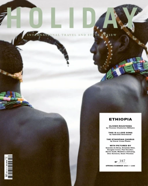 Magazine cover featuring two men with dark skin, adorned with traditional tribal accessories, against a blurred white background. The magazine is titled HOLIDAY.