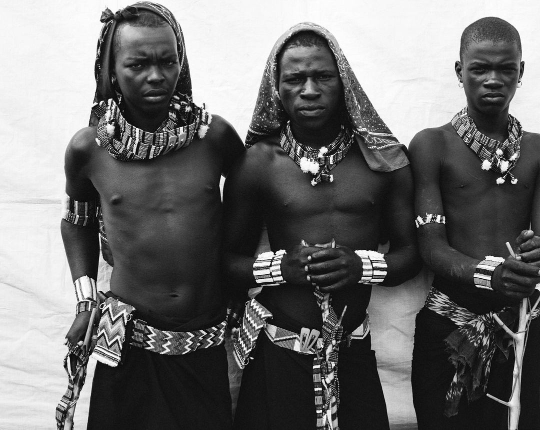 Three African men stand shoulder-to-shoulder wearing traditional beaded jewellery, cloth wraps and skirts, against a white backdrop.