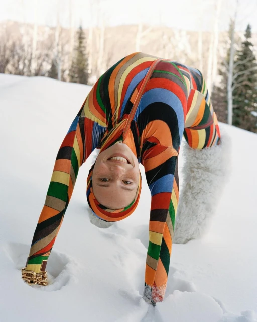A woman in a colorful patterned snowsuit bends over backwards in the snow with a snowy forest visible in the background on a bright day.
