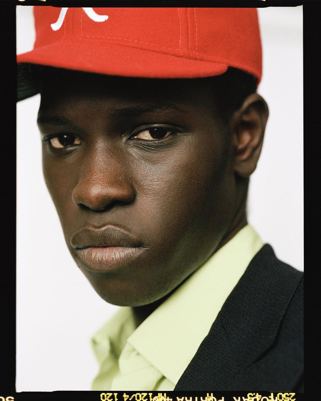 Close-up portrait of a young Black man wearing a red and white baseball cap, buttoned green shirt, and black suit jacket against a white background.