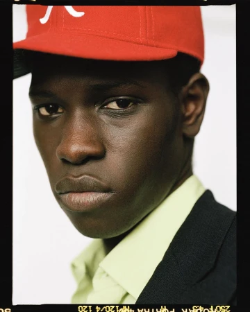 Close-up portrait of a young Black man wearing a red and white baseball cap, buttoned green shirt, and black suit jacket against a white background.