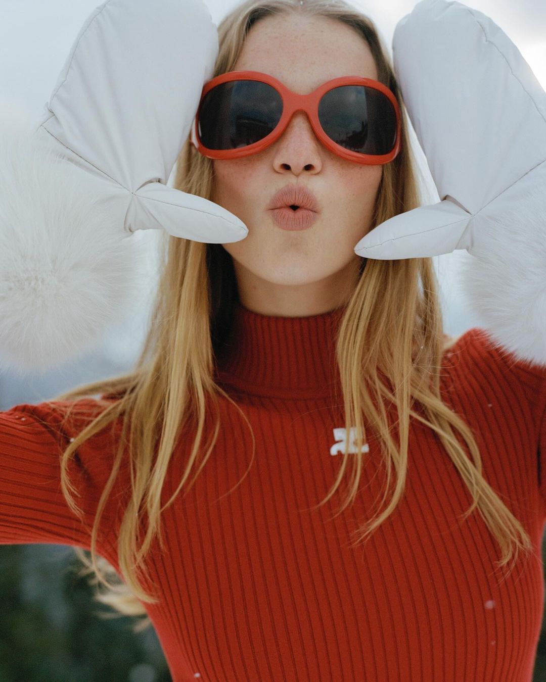 A blond woman in an orange turtleneck and orange sunglasses makes a face with white mittens near her head in a snowy environment.