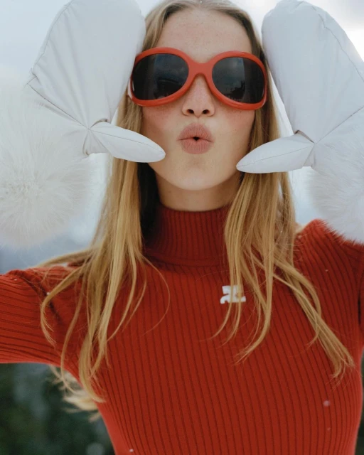A blond woman in an orange turtleneck and orange sunglasses makes a face with white mittens near her head in a snowy environment.