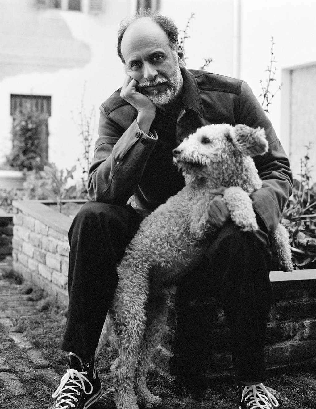 Black and white portrait of a bearded man holding a fluffy dog while seated outdoors near low brick walls
