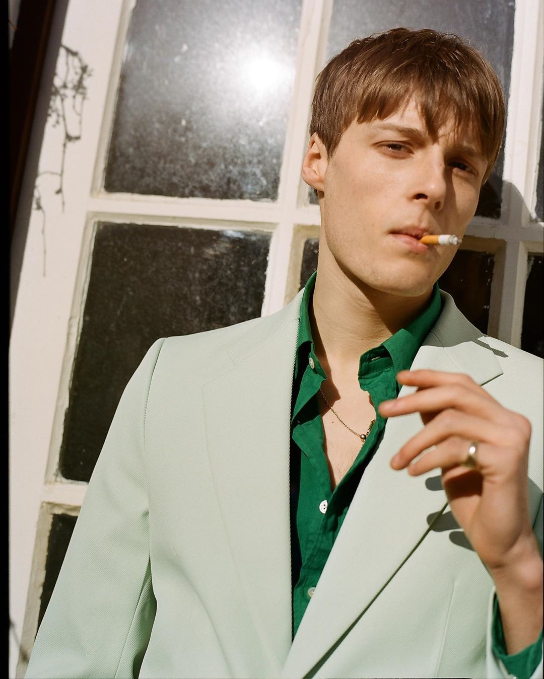 Young man in a light green suit and green shirt smoking a cigarette, standing in front of a white window frame with natural light.