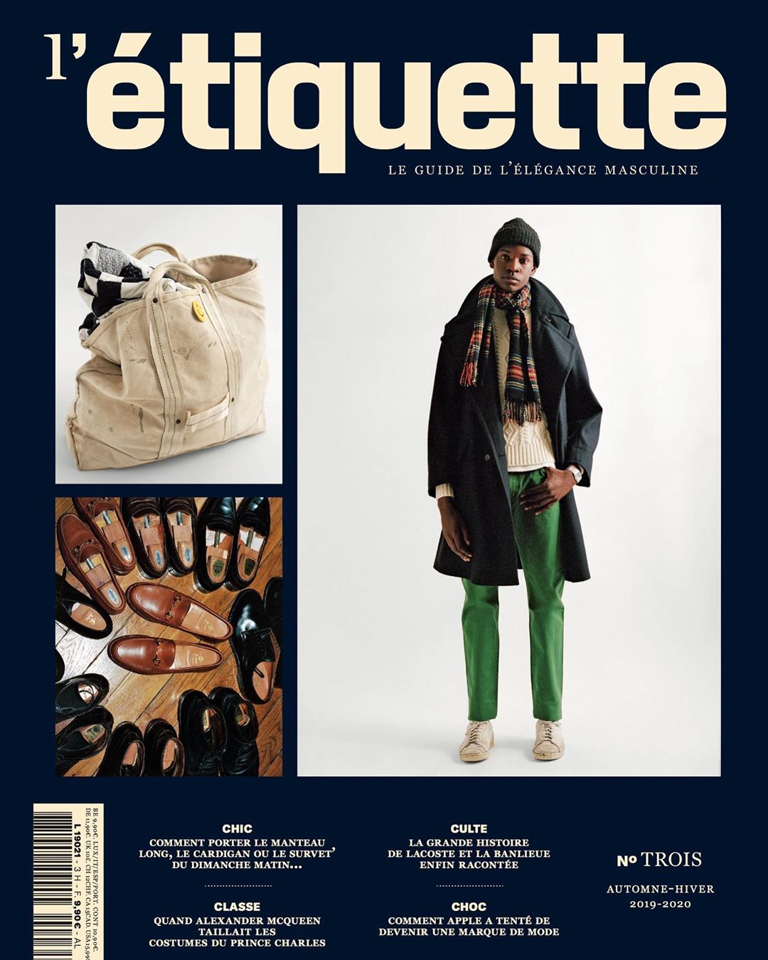 A magazine cover with a man in a dark coat and green pants, a travel bag, and shoes, set against a navy blue background.