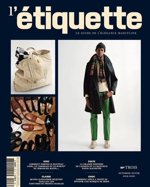 A magazine cover with a man in a dark coat and green pants, a travel bag, and shoes, set against a navy blue background.