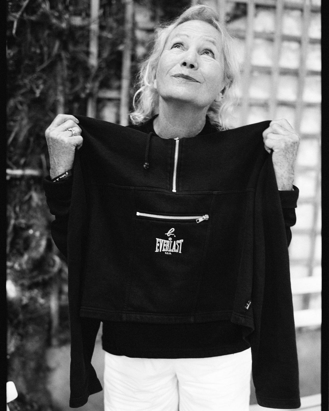 Black-and-white portrait of an elderly woman holding up a black Everlast sweatshirt against a background of greenery and a fence.