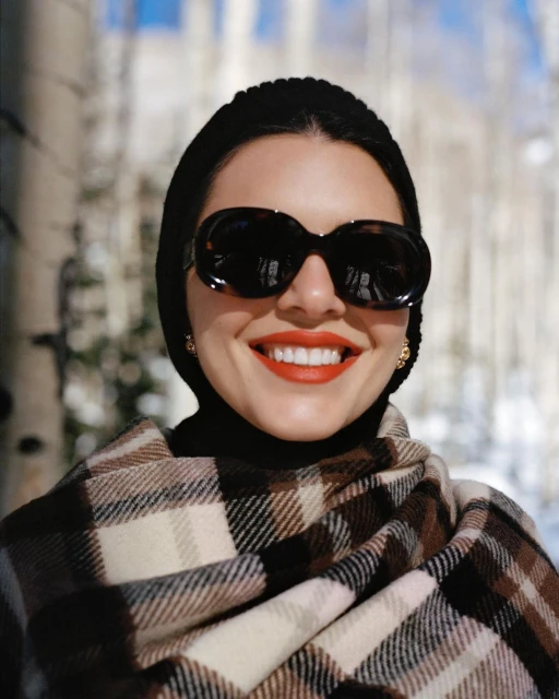 Portrait of a smiling woman in sunglasses and a head covering, wrapped in a plaid shawl, with birch trees in the blurred background.