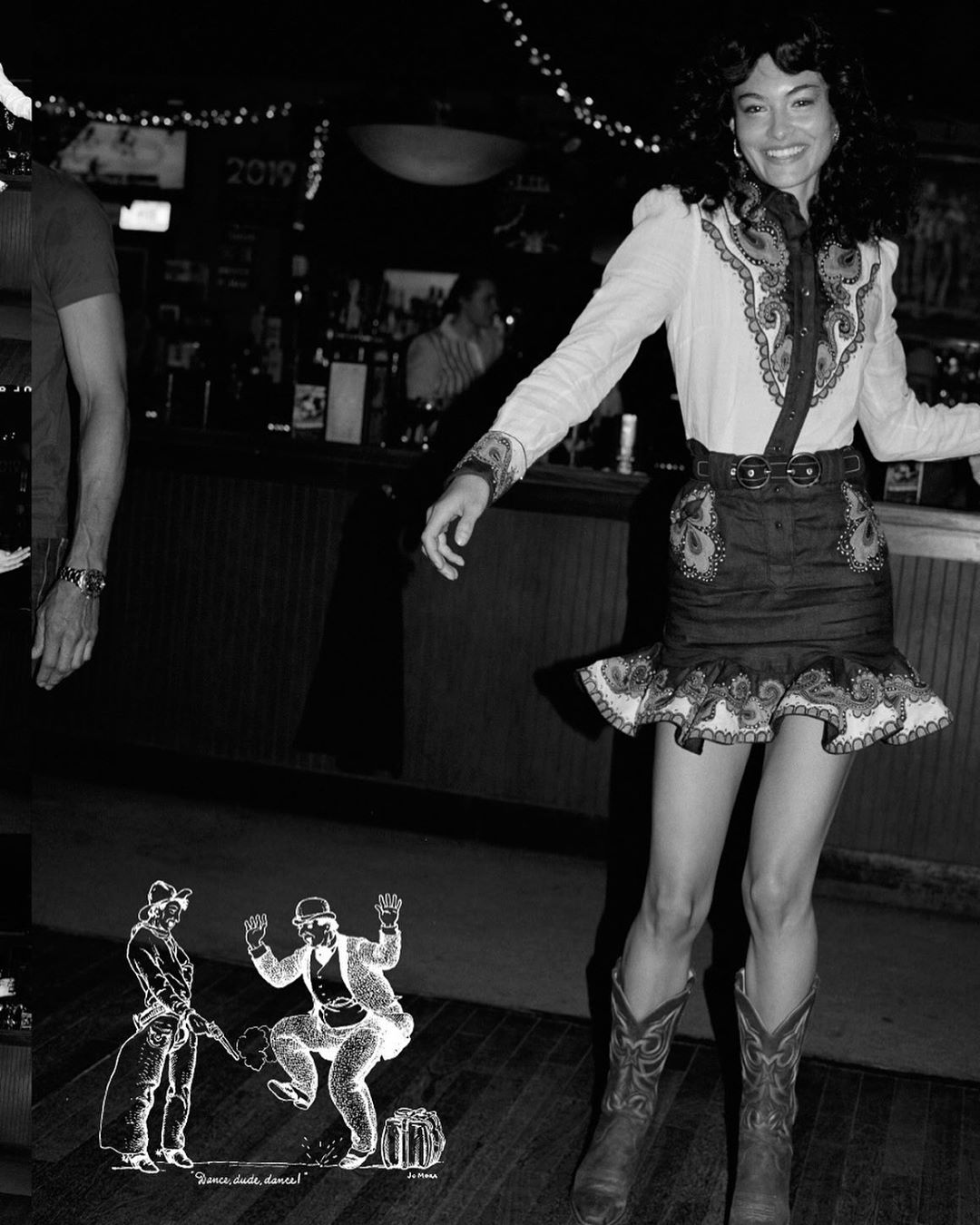 A woman smiles, wearing a western-style blouse, skirt, and cowboy boots, standing on a dance floor in a dimly lit, busy bar.
