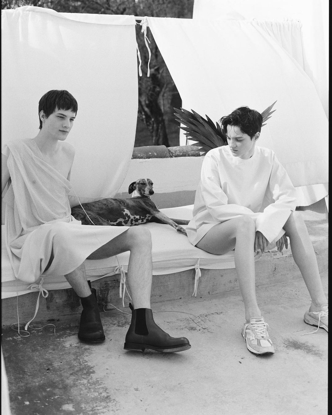 Two men sit outside a white tent-like structure with a dog, one wearing a draped garment and boots, the other casual shirt and sneakers.