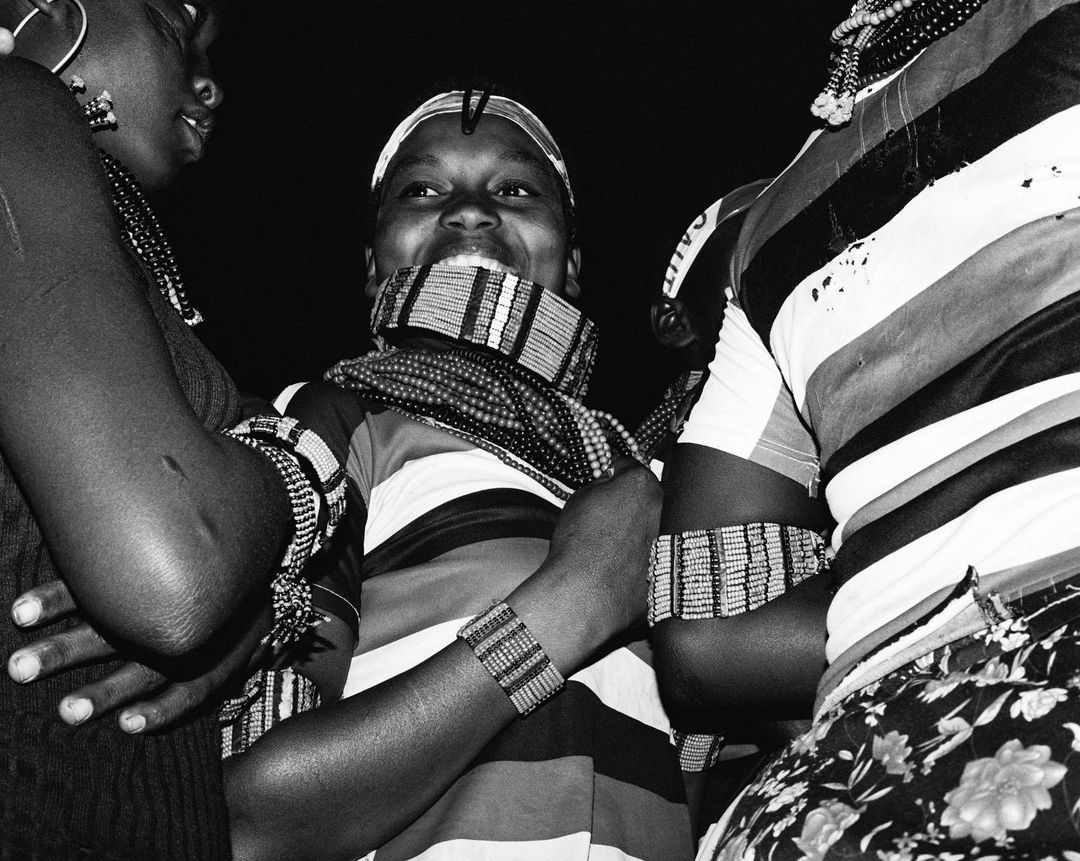 Three women with beaded jewelry and striped clothing, captured in black and white, showcasing cultural adornments and close social interaction.
