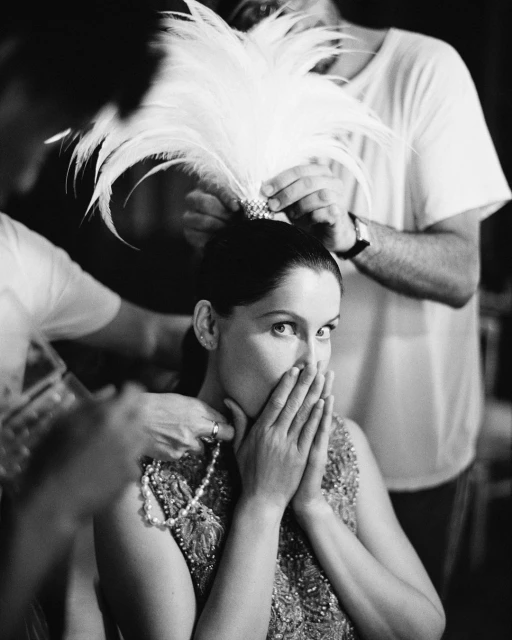 Elegant woman with feather headdress and beaded dress being styled in photo studio by two stylists, captured in black and white.