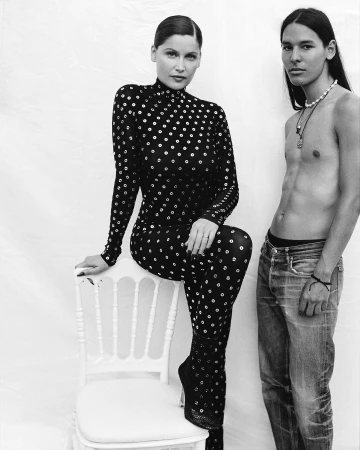 Black and white portrait of a woman in a patterned catsuit seated on a white chair next to a shirtless man in jeans.