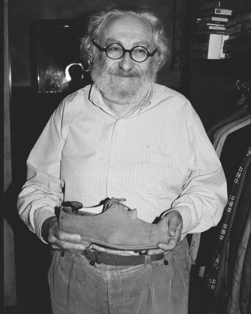 Portrait of an elderly man with a beard, holding a suede shoe indoors, with clothing racks in the background, shot in black and white.
