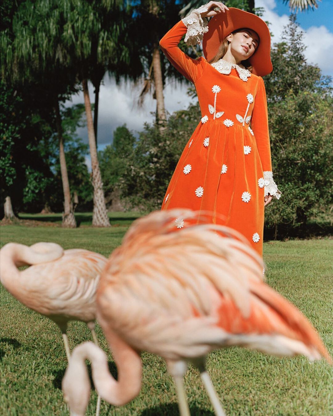 Elegant woman in an orange dress and hat poses with pink flamingos in a lush green outdoor setting, fashion-forward and stylish.