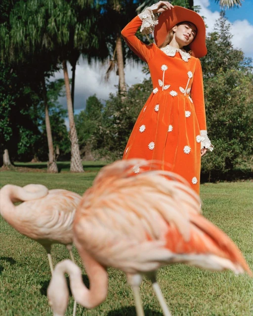 Elegant woman in an orange dress and hat poses with pink flamingos in a lush green outdoor setting, fashion-forward and stylish.