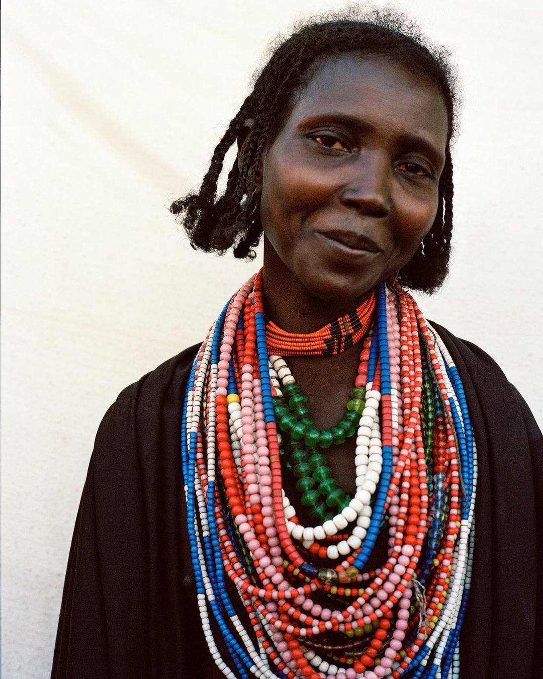 Portrait of a woman wearing layered colorful beaded necklaces and dark garments against a plain white background.