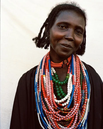 Portrait of a woman wearing layered colorful beaded necklaces and dark garments against a plain white background.