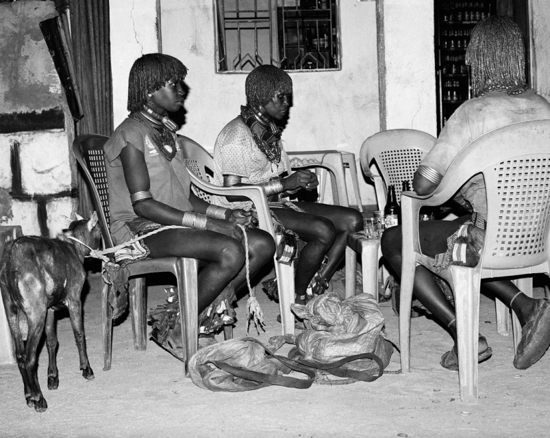 Three traditionally dressed adult males sit on plastic chairs indoors with a goat nearby. Black and white image of people in native garb.