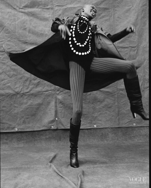 A dark-skinned woman in motion wearing a black coat, top, patterned tights, boots, and a pearl necklace poses in front of a gray backdrop.