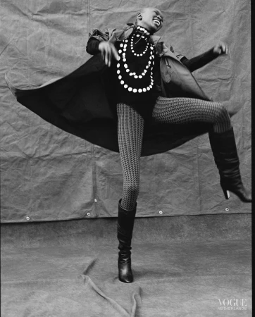 A dark-skinned woman in motion wearing a black coat, top, patterned tights, boots, and a pearl necklace poses in front of a gray backdrop.
