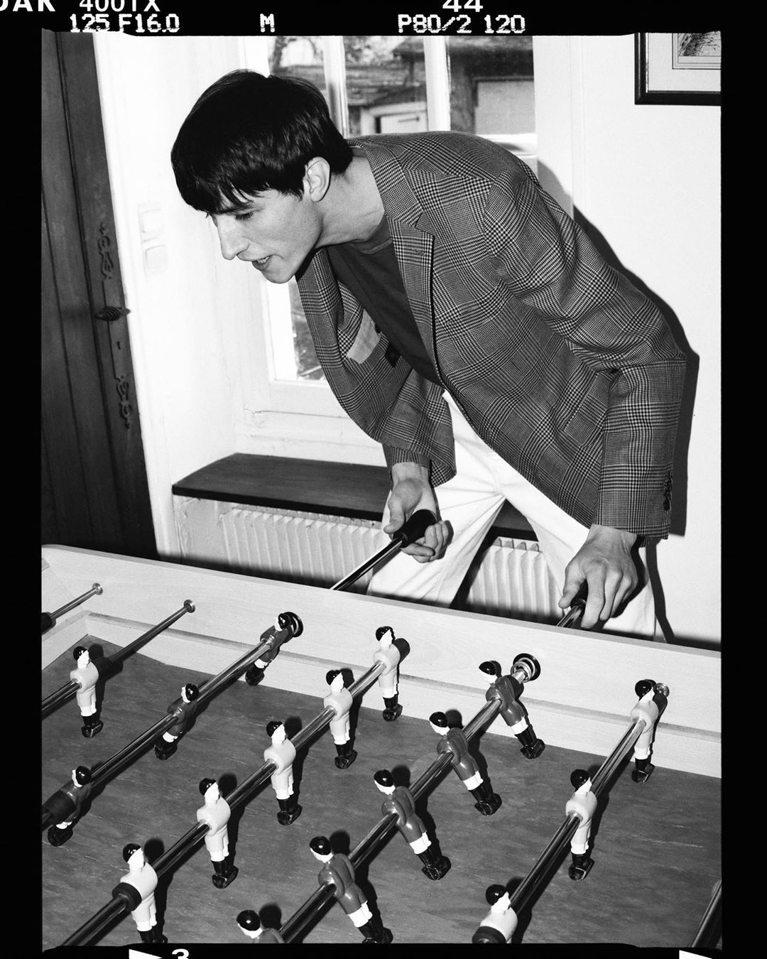 Monochrome indoor shot of a male playing foosball, wearing a patterned blazer and white pants, in a naturally lit room.