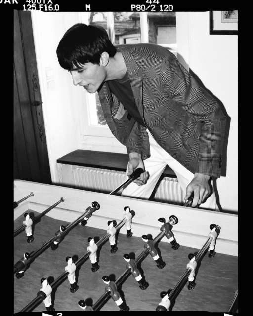 Monochrome indoor shot of a male playing foosball, wearing a patterned blazer and white pants, in a naturally lit room.