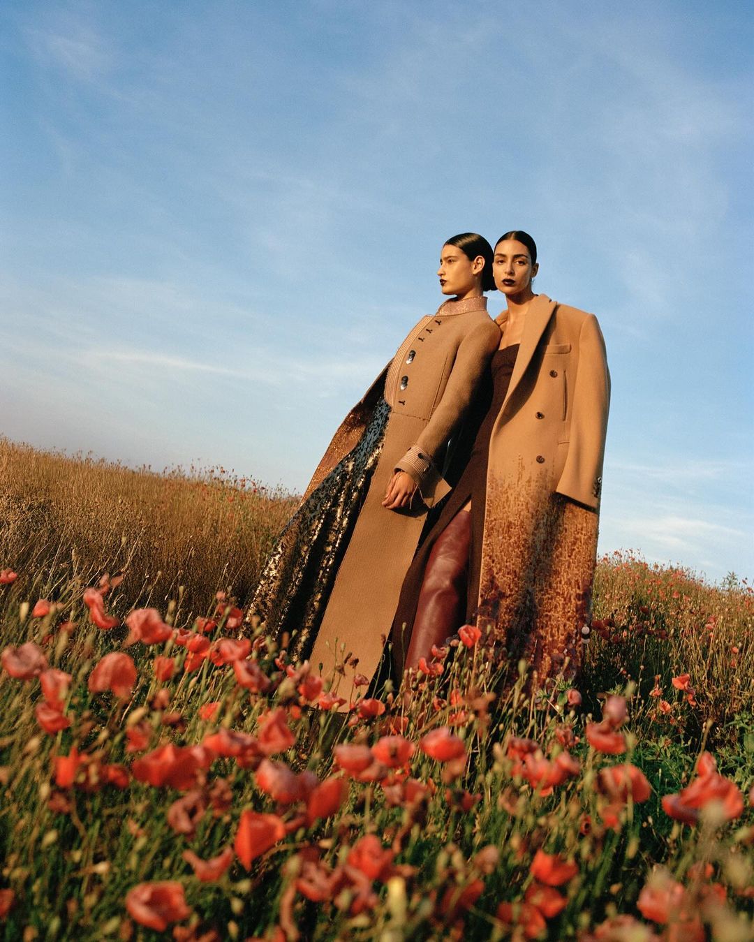 Two women pose in tan coats amongst red flowers under a blue sky, fashion editorial, outdoor setting, portrait photography.