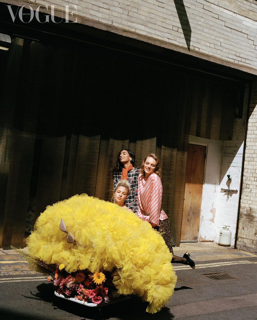 Three fashion models in an urban alleyway, one riding in a repurposed shopping cart filled with flowers and a bright yellow ruffled tulle.