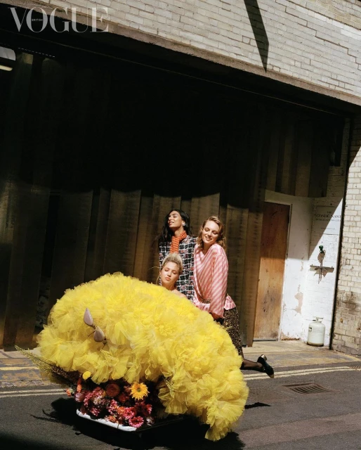 Three fashion models in an urban alleyway, one riding in a repurposed shopping cart filled with flowers and a bright yellow ruffled tulle.