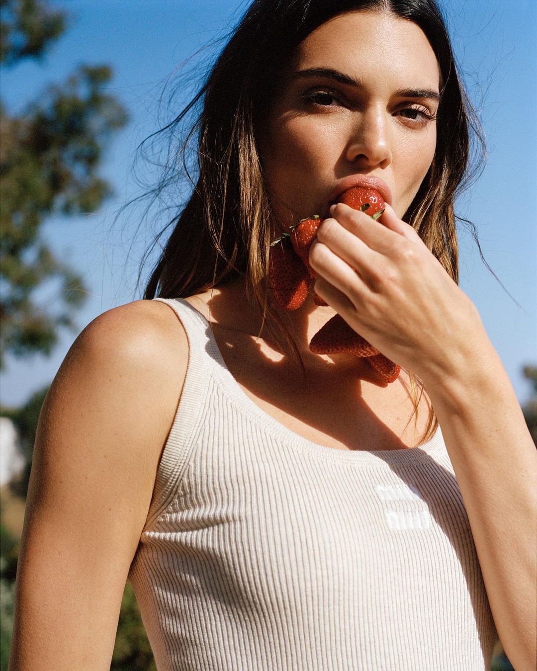 Close-up of a woman eating strawberries outdoors on a sunny day, wearing a white ribbed tank top, with greenery in the background.