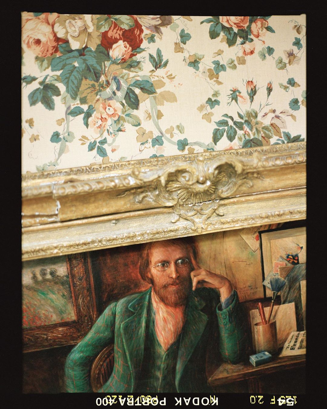 Painting of a seated man in a green suit against a floral wallpaper backdrop, framed in gold, vintage aesthetic, portrait.