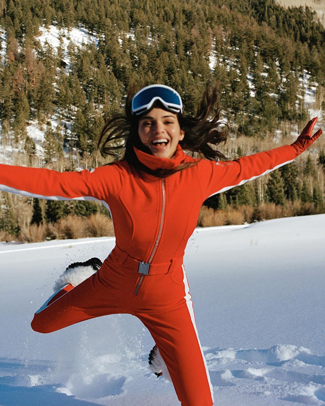 Woman in a red ski suit jumps with joy on a snowy day in the mountains near a pine forest. She is wearing ski goggles.