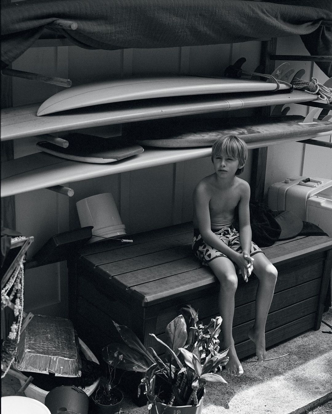 Black and white portrait of a shirtless young boy sitting on a bench with surfboards on a rack behind him in an indoor setting.