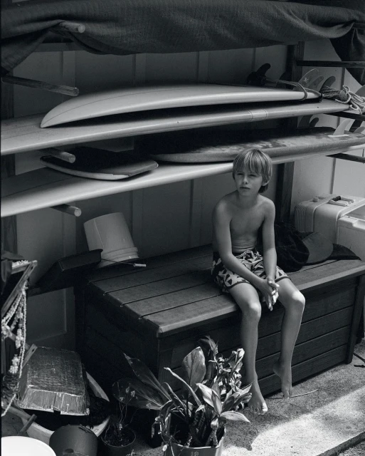 Black and white portrait of a shirtless young boy sitting on a bench with surfboards on a rack behind him in an indoor setting.