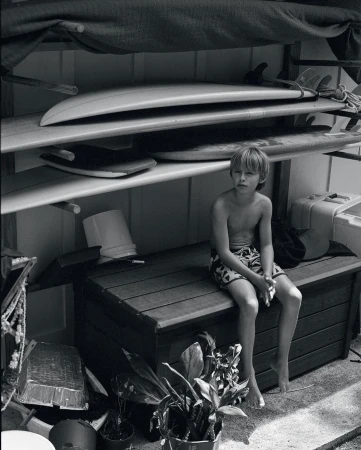 Black and white portrait of a shirtless young boy sitting on a bench with surfboards on a rack behind him in an indoor setting.