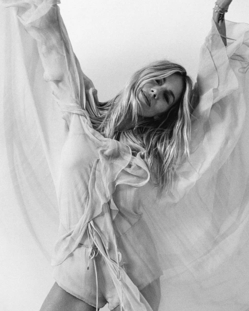 Black and white portrait of a blonde woman in a flowing garment raising her arms in a simple indoor setting.