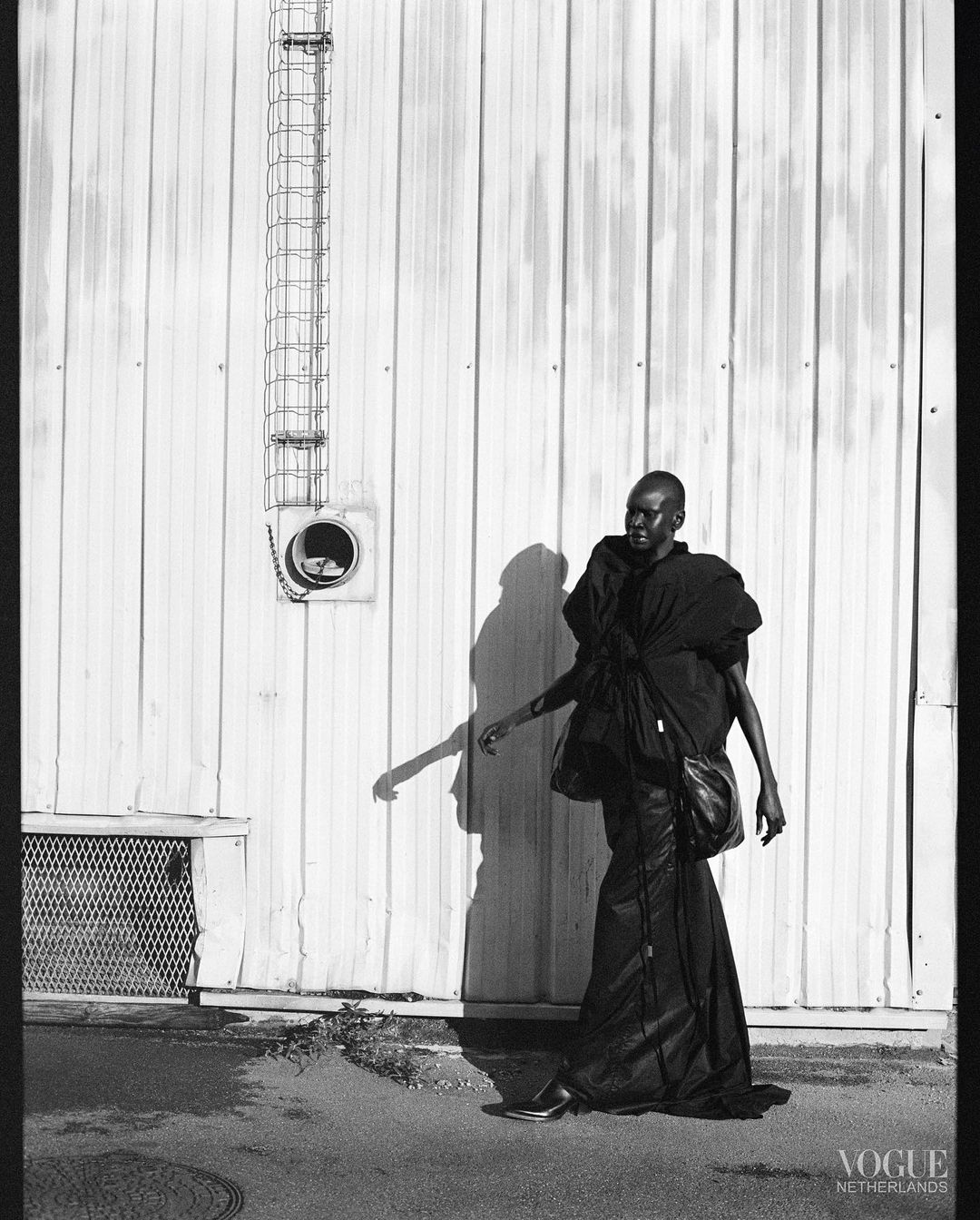 Black and white fashion photograph of a dark-skinned adult model in an oversized black outfit standing against a weathered industrial wall.