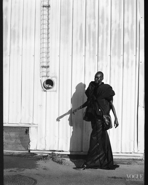 Black and white fashion photograph of a dark-skinned adult model in an oversized black outfit standing against a weathered industrial wall.