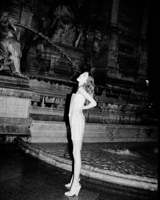 A full shot of a woman standing in front of a decorative fountain with her head tilted back looking up at the water falling down.