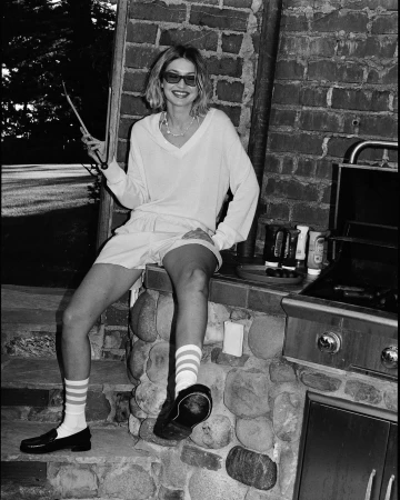 Blonde woman in casual outfit smiles while sitting on a stone counter with a grill and cooking utensils outdoors in black and white.