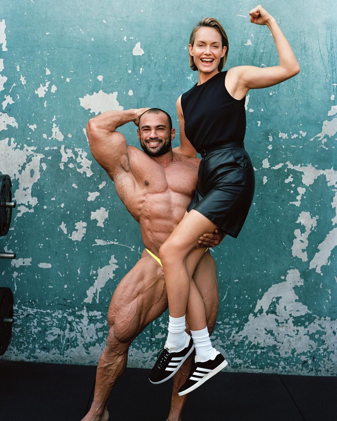 A muscular bodybuilder holds a smiling woman in his arms against a textured aqua wall, showcasing physique and camaraderie.
