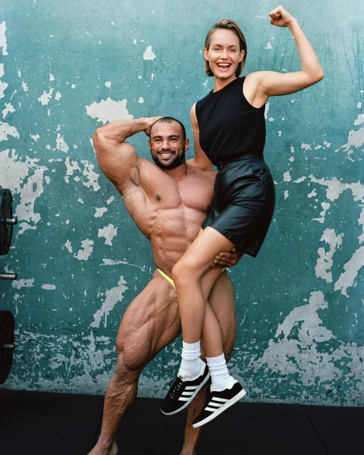 A muscular bodybuilder holds a smiling woman in his arms against a textured aqua wall, showcasing physique and camaraderie.
