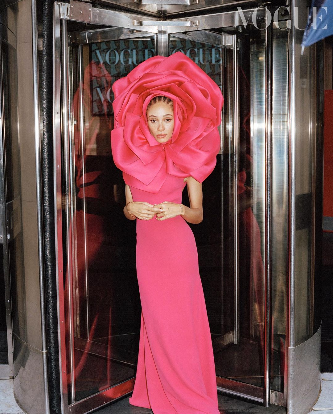 Woman in a pink dress with a large pink flower headpiece, standing in front of revolving doors with Vogue magazine covers visible.